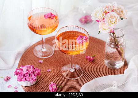 Deux cocktails décorés de fleurs sur une table blanche à côté de la fenêtre et un vase avec des roses en arrière-plan. Banque D'Images