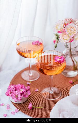 Deux cocktails décorés de fleurs sur une table blanche à côté de la fenêtre et un vase avec des roses en arrière-plan. Banque D'Images