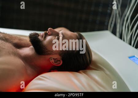 Gros plan sur le beau jeune homme allongé sur un lit de massage électrique et bénéficiant d'un soin spa dans un salon de spa de luxe. Musclé mâle ayant le repos Banque D'Images