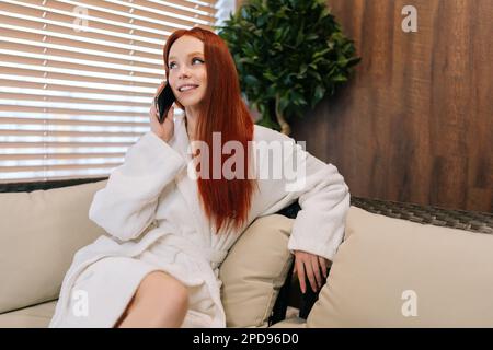 Portrait d'une charmante jeune femme à tête rouge portant un peignoir blanc, assise sur un canapé confortable près de la fenêtre et parlant sur un smartphone, en appréciant la conversation Banque D'Images