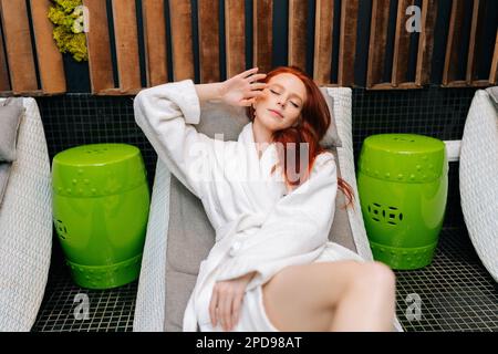 Portrait d'une jolie jeune femme à tête rouge portant un peignoir blanc sur le visage allongé avec des yeux fermés sur un transat après un soin spa au salon de beauté. Banque D'Images