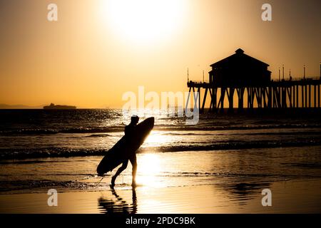 Silhouette d'un surfeur marchant sur la plage transportant une planche de surf devant la jetée de Huntington Beach, Californie au coucher du soleil Banque D'Images