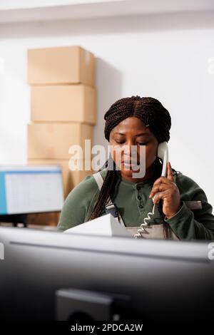 Le superviseur de la réserve se tient au comptoir et parle avec les clients distants au moyen d'un téléphone fixe pour discuter des détails de la commande. Employé afro-américain travaillant à l'inventaire des marchandises dans l'entrepôt Banque D'Images