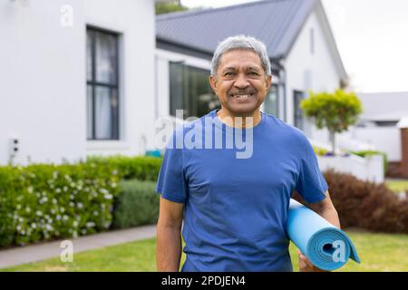 Portrait d'homme biracial senior souriant portant un tapis de yoga et debout contre la maison dans la cour Banque D'Images