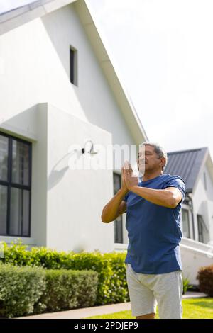 Vue à angle bas de l'homme aîné biracial méditant dans la pose de prière tout en se tenant contre la maison dans la cour Banque D'Images