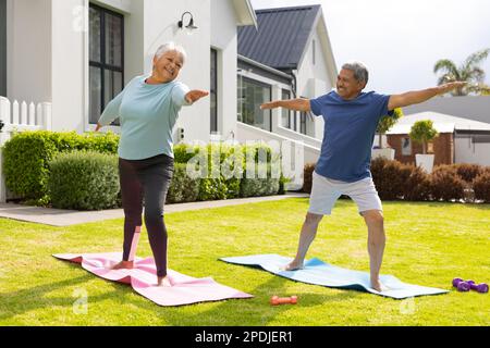 Couple biracial senior souriant avec bras surtendus guerrier pratiquant 2 pose sur des tapis dans la cour Banque D'Images