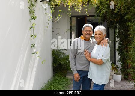 Portrait d'un couple aîné biracial aimant souriant et debout près des plantes dans la cour contre la maison Banque D'Images
