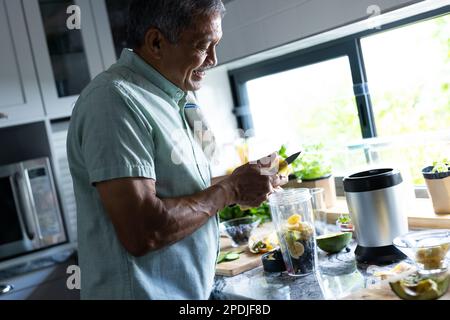 Vue latérale d'un homme biracial souriant coupant des tranches de banane dans un mélangeur sur le comptoir de la cuisine Banque D'Images