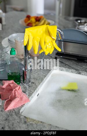 Distributeur de savon par gants de lavage jaunes sur le robinet et l'évier de cuisine rempli d'eau savonneuse Banque D'Images