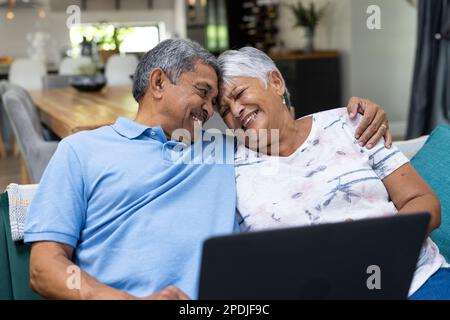 Joyeux couple biracial senior avec tête à tête en utilisant un ordinateur portable tout en se relaxant sur le canapé Banque D'Images