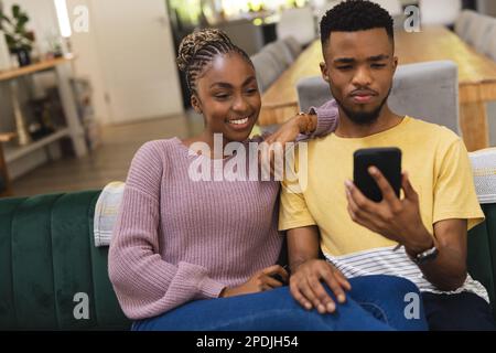 Heureux couple afro-américain assis sur un canapé et utilisant un smartphone Banque D'Images