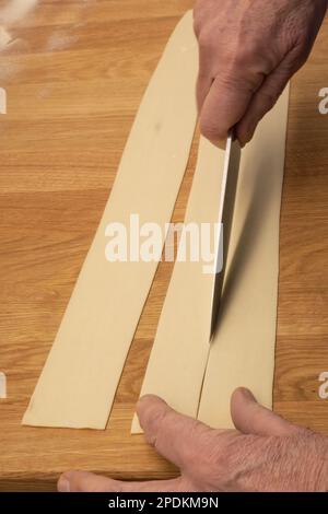 Production de pâtes faites maison - nouilles italiennes à faire soi-même - gros plan des mains qui coupent la pâte enroulée pour préparer des pâtes faites soi-même Banque D'Images