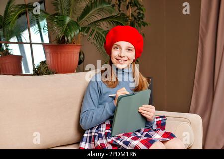 Une adolescente tire sur une tablette numérique dans le salon. Portrait gaie écolière fille en béret rouge. Concept d'éducation. Banque D'Images