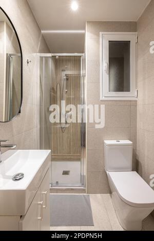 Salle de bains moderne et compacte avec carrelage beige, cabine de douche en verre, lavabo et miroir, toilettes blanches avec fenêtre. Concept d'une petite salle de bains dans un hôtel ou Banque D'Images