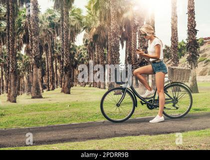 Jeune fille blonde avec de longs cheveux est sur un bycicle et de prendre une photo d'une belle allée de palmiers. Banque D'Images