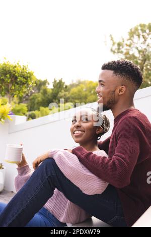 Heureux couple afro-américain assis sur les escaliers dans le jardin, boire du café et embrasser Banque D'Images