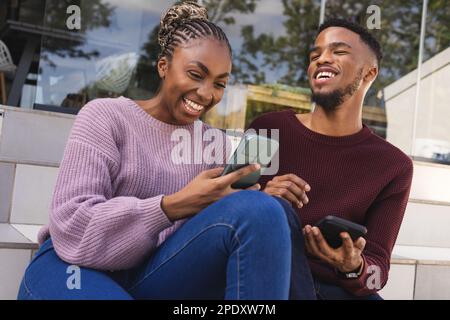 Heureux couple afro-américain assis sur les escaliers dans le jardin, en utilisant des smartphones Banque D'Images
