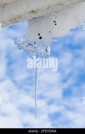 La glace transparente est suspendue sur un gouttière blanche d'une maison rurale sous un ciel nuageux par un beau jour de printemps Banque D'Images