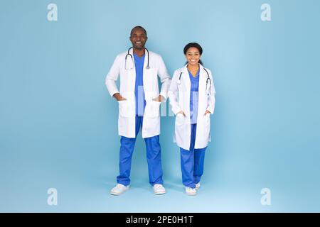 Afro-américain hommes et femmes médecins posant vêtu d'uniforme blanc sur fond bleu, prise de vue en studio, pleine longueur Banque D'Images