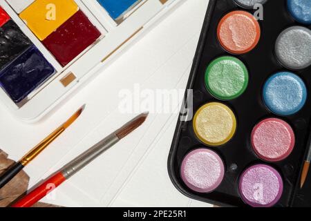 Gros plan sur les fournitures artistiques : peintures à l'aquarelle régulières et brillantes, pinceaux, papier. 2 jeux d'aquarelles préparés pour un artiste Banque D'Images