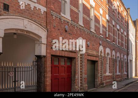 Un bâtiment en brique avec fenêtres voûtées dans une rue arrière. Une entrée voûtée se trouve au-dessus d'une porte en fer ornée. Le soleil s'illumine à l'étage supérieur. Banque D'Images