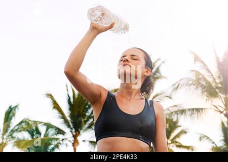 Femme athlète versant de l'eau sur son visage de la bouteille après entraînement dur dans le climat tropical. Banque D'Images