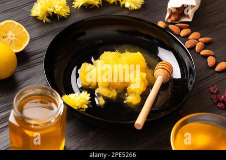 Miel cristallisé, balancier dans une assiette noire sur fond en bois foncé. Composition de miel, de citron, de fleurs jaunes et de pétales sur fond noir. Banque D'Images