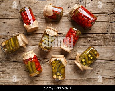 Conserves de concombres et de tomates en pots de verre sur un fond en bois ancien vue du dessus. Tomates cerises en conserve et carnishons en pots avec couvercles kraft. Banque D'Images