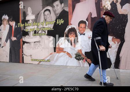 Un homme handicapé marche avec l'aide de béquilles devant un palissade de détail mettant en vedette une jeune mariée et un couple marié célébrant leur mariage, une image de loyauté, le dévouement de la jeunesse à l'âge de la retraite, le 13th mars 2023, à Londres, en Angleterre. Banque D'Images