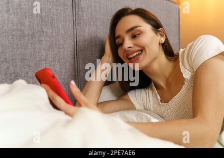 Une belle jeune fille se trouve dans le lit et aime la communication, tient un smartphone dans ses mains. Banque D'Images