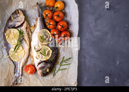 Savoureux poisson entier cuit sur du papier de cuisson. Dorade cuite au four avec citron, oignon, herbes, tomates cerises, épices sur fond rustique foncé. Grillé Banque D'Images