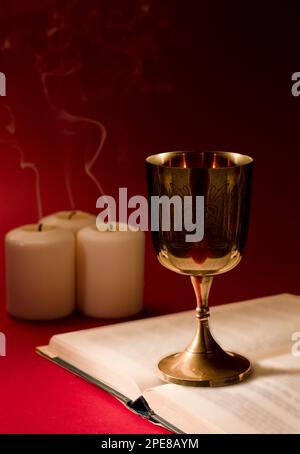 le calice chrétien sur la bible ouverte avec des bougies fumeuses et un arrière-plan rouge fort Banque D'Images