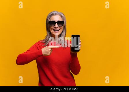 Portrait d'une femme aînée souriante montrant ou présentant une application de téléphone mobile et pointant le doigt vers l'écran du smartphone à portée de main. Femme mature isolée sur fond jaune Banque D'Images