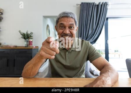 Portrait d'un homme biracial chevronné gai pointant vers la caméra tout en parlant par vidéoconférence à table Banque D'Images