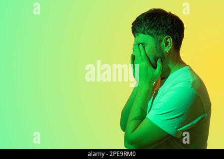 Portrait créatif d'un homme d'âge moyen barbu. L'homme déprimé avec des problèmes mentaux couvre son visage avec ses mains et pleure. Lumière verte colorée Banque D'Images