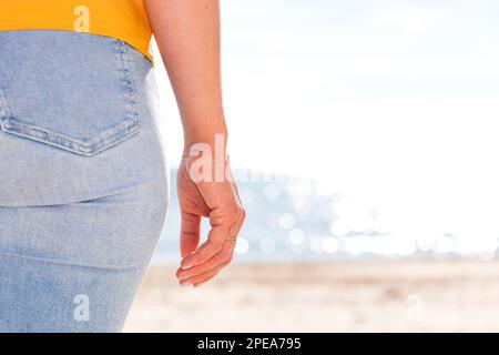 Anonyme femme en chemise jaune et jeans en denim debout sur une plage de sable près de la mer ondulée le jour ensoleillé pendant les vacances d'été Banque D'Images