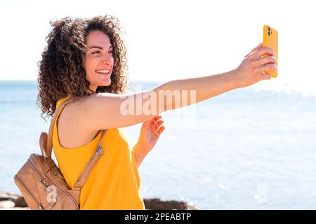 Jeune femme gaie avec des cheveux bouclés dans une tenue décontractée avec sac à dos souriant et prenant autoportrait sur le smartphone tout en se tenant debout sur la mer pendant Banque D'Images