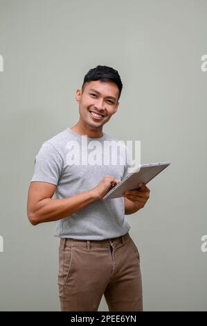 Beau et souriant homme asiatique du millénaire dans des vêtements décontractés tenant une tablette numérique et debout sur un fond vert isolé. Banque D'Images