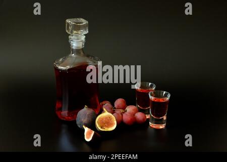 Un décanter en verre antique et deux verres de liqueur maison sur fond noir, à côté de morceaux de figues mûres et d'un bouquet de raisins rouges. Gros plan. Banque D'Images