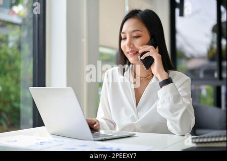 Femme d'affaires asiatique du millénaire attrayante et professionnelle parlant au téléphone avec son partenaire d'affaires tout en travaillant dans son bureau. Banque D'Images