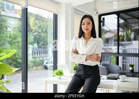 Femme d'affaires ou PDG asiatique du millénaire confiante et prospère, vêtue de vêtements décontractés, assis sur une table avec les bras croisés au bureau. Banque D'Images