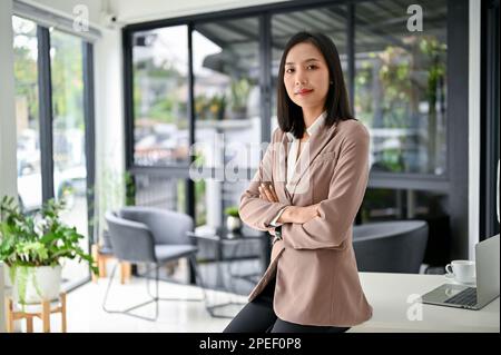 Une femme d'affaires ou PDG asiatique confiante et prospère du millénaire en costume d'affaires formel est assise sur une table dans son bureau moderne avec des bras croisés. Banque D'Images