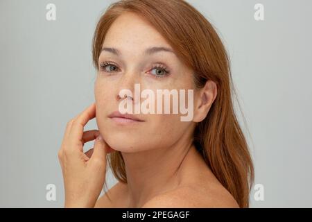 Portrait d'une femme d'âge moyen avec cheveux rougeâtres tenant la main sur la joue regardant l'appareil photo sur fond gris Banque D'Images