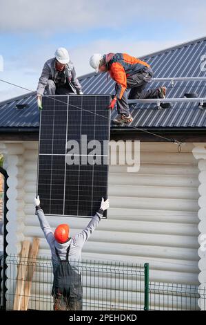 Techniciens professionnels soulevant le panneau solaire sur un toit de maison. Trois travailleurs en halmets et combinaisons de travail transportant une cellule solaire lourde. Installation de la batterie PV. Banque D'Images