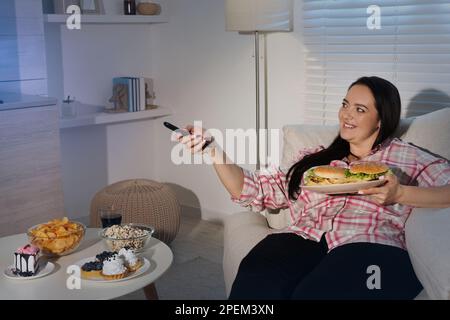 Bonne femme en surpoids avec de la nourriture malsaine regardant la télévision sur le canapé à la maison Banque D'Images