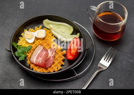 Poivre frais et coin de tomate, gaufres, bacon frit et œufs dans une poêle. Une tasse de thé et une fourchette sur la table. Banque D'Images