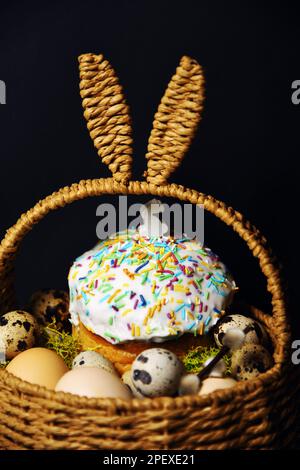 Décorations de Pâques dans un panier avec oreilles de lapin. Gâteau de Pâques et œufs sur mousse, œufs de caille et plumes sur fond noir foncé. Une petite figure d'un Banque D'Images