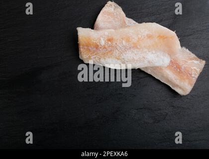 Poisson congelé sur une assiette noire, filet de morue blanc, filet de merlu glacé, viande de goberge congelée sur fond sombre vue de dessus Banque D'Images