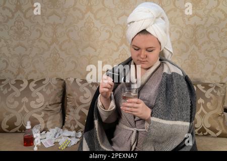 Une fille qui présente les symptômes d'un rhume coule un médicament d'une bouteille dans un verre d'eau. Concept de médecine, de soins de santé, de pharmacie et de traitement à domicile Banque D'Images