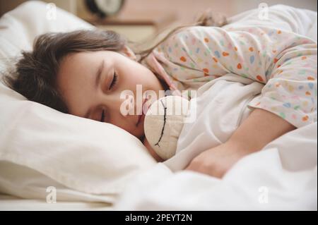 Portrait en gros plan d'une petite fille mignonne qui embrasse doucement son jouet tout en dormant dans le lit dans la chambre à coucher claire Banque D'Images